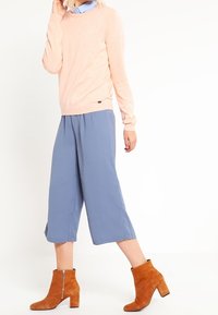 Pull rose clair, culottes bleues et bottines brunes. Le pull a un col rond ; les pantalons sont larges. Les bottines sont dotées d'une fermeture éclair sur le côté.