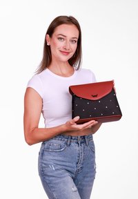 Černá taška s růžovými puntíky, červený kožený klapka, obdélníkový tvar, hladký povrch a detail loga na přední straně.