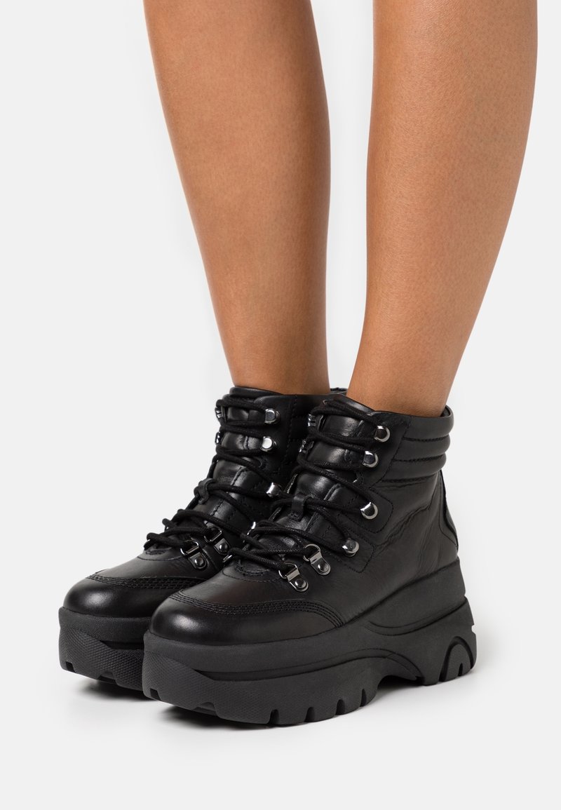 Bottes noires à lacets jusqu'à la cheville avec une semelle épaisse, cuir texturé, col rembourré et œillets métalliques pour les lacets. Design avec plateforme légèrement surélevée.