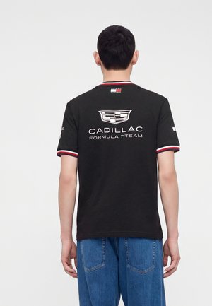 Homme de dos portant un t-shirt noir de l’équipe Cadillac Formula 1 avec des bordures rouges, blanches et bleues et un jean bleu sur un fond uni.