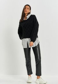 Schwarzer gehäkelter Pullover mit einem strukturierten Muster, übereinander getragen mit einem weiß-schwarz gestreiften Hemd mit Knöpfen. Kombiniert mit schwarzen Lederhosen und weißen Turnschuhen.
