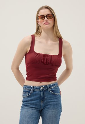 Donna che indossa occhiali da sole con lenti color ambra, un top senza maniche bordeaux arricciato e jeans blu decorati con piccole perle.