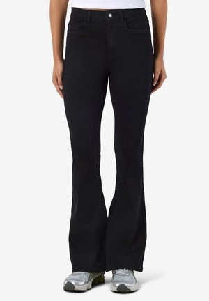 Femme portant un pantalon évasé taille haute noir, un top court blanc et des baskets argentées, debout les mains détendues le long du corps.