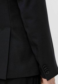 Czarna marynarka z teksturowanej tkaniny, ostre klapy i szczegółami czterech guzików na mankiecie. Dopasowany krój z rozcięciem z tyłu.