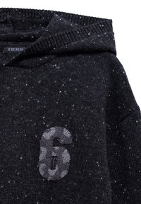 Felpa nera a maglia con una texture a macchie, dotata di una patch in feltro grigia con il numero "6" sul davanti. Maglia a coste attorno al collo e al cappuccio.