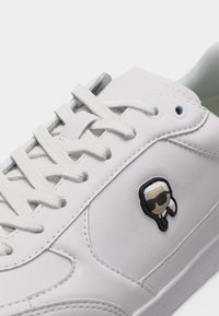 Λευκό αθλητικό παπούτσι με λευκά κορδόνια και μικρό λογότυπο καρτούν χαρακτήρα που φοράει γυαλιά ηλίου στην πλαϊνή πλευρά.