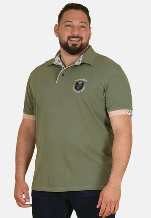 Homme souriant portant un polo vert olive avec un col et des poignets floraux, pantalon foncé, debout devant un fond uni.