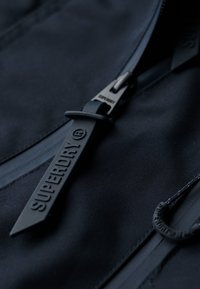 Zwarte jas met een matte ritssluiting met het opschrift "SUPERDRY" in reliëf. Gladde stof, minimale textuur en een strak ontwerp zonder zichtbare patronen.