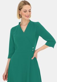 Zielona sukienka w kopertowym kroju z dekoltem w serek, rękawami o długości trzech czwartych oraz ozdobnymi guzikami w talii. Wykonana z lekkiego, strukturalnego materiału.