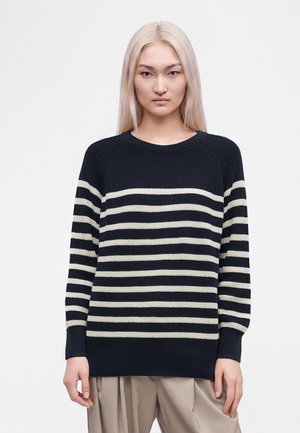 Jonge vrouw met lang blond haar, die een zwart-wit gestreepte trui en beige broek draagt, staand tegen een effen witte achtergrond.