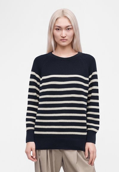 Jeune femme aux longs cheveux blonds portant un pull rayé noir et blanc et un pantalon beige, debout devant un fond blanc uni.