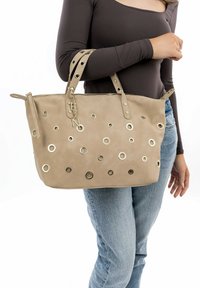 Bolso beige de ante sintético, con adornos metálicos circulares y cierre lateral con cremallera. Dos asas con detalles de tachuelas.