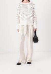 Woman wearing a white knit sweater and wide-leg cream trousers, holding a small black handbag, standing against a white background.