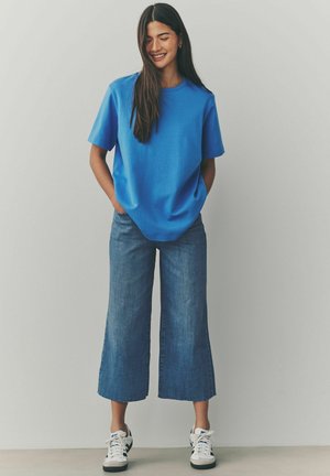 Blaue Baumwoll-T-Shirt mit kurzen Ärmeln, kombiniert mit weit geschnittenen Jeanshosen. Das Model trägt weiße Sneakers mit schwarzen Akzenten.