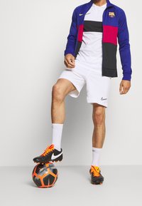 Blue and red zip-up jacket with a logo, white T-shirt, white shorts, and black football boots with orange accents, standing on a patterned ball.