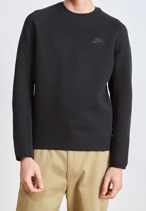 Hombre vistiendo sudadera negra de Nike con mangas largas y pantalones beige, de pie contra un fondo claro y sencillo.