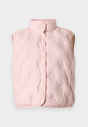 Femme portant un gilet matelassé rose sur un top noir rayé, un jean bleu à jambes larges et des baskets beige, debout avec une main dans la poche.