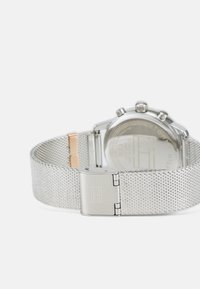 Montre en maille argentée avec un boîtier en acier inoxydable, une boucle polie et un cadran rond visible à l'arrière, avec une gravure de la marque.