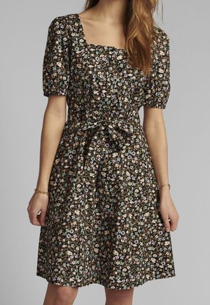 Robe noire à motifs floraux avec manches courtes bouffantes, encolure carrée et ceinture nouée à la taille, portée par une femme aux longs cheveux châtain clair.