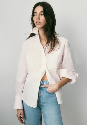 Hosszú sötét hajú nő, aki világos rózsaszín, túlméretezett inget és kék farmert visel, egyik kezét a zsebében tartva, balra néz egy egyszínű háttér előtt.