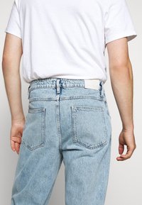 Jeans de mezclilla azul claro con una cintura alta, bolsillos traseros y una cinturilla elástica. Combinados con una camiseta blanca, se centra en la vista trasera de la prenda.