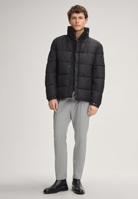 Giacca a vento nera con colletto alto e bottoni a pressione, indossata sopra una camicia nera, abbinata a pantaloni grigi chiari e stivali neri.