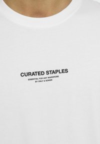 Weißes Baumwoll-T-Shirt mit schwarzem Text, der "CURATED STAPLES" und "ESSENTIAL FOR ANY WARDROBE" in einer schlichten, modernen Schriftart zeigt.