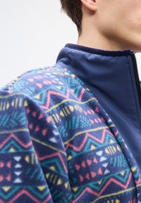 Φλις μπουφάν με ζωηρό γεωμετρικό σχέδιο σε μπλε, ροζ, κίτρινο και πράσινο. Ναυτικό γιακά και ολόκληρο φερμουάρ. Μαλακή υφή.