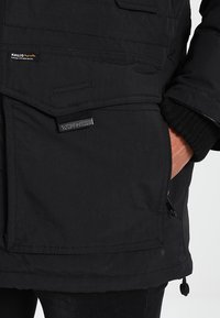 Poche de veste noire avec rabat et fermeture éclair, main partiellement insérée dans la poche latérale, étiquettes de marque KHUJO visibles sur le tissu.