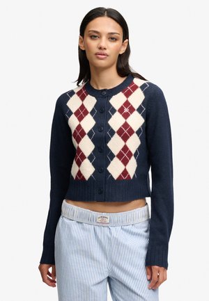 Femme portant un cardigan bleu marine boutonné avec un motif losanges rouge et blanc et un pantalon rayé bleu clair et blanc avec une étiquette Superdry.
