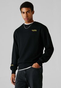 Mladý muž v čiernej mikine Red Bull Racing, s strieborným retiazkou a tmavými rifľami, stojaci s jednou rukou v vrecku na šedom pozadí.