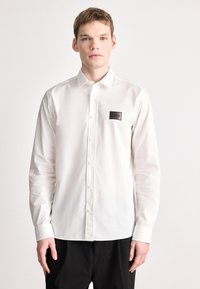 Λευκό πουκάμισο με μακριά μανίκια και γιακά με κουμπιά, με μαύρο επώνυμο ταμπελάκι στο αριστερό στήθος και απαλό βαμβακερό ύφασμα.