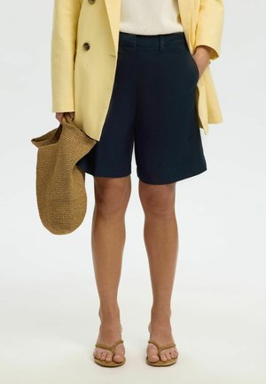 Persona con pantalones cortos azul marino hasta la rodilla, blazer amarillo, top beige, sandalias marrones y sosteniendo un gran bolso marrón tejido.