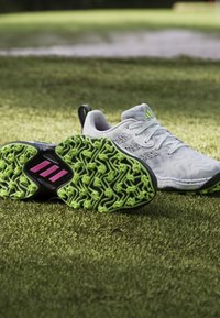 White sports shoes with a textured upper, featuring green and black rubber soles. Notable pink accents on the sole’s design.