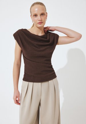 Žena v bezrukávovom tmavohnedom štruktúrovanom topu a béžových plisovaných širokých nohaviciach stojí na jednoduchom bielom pozadí.