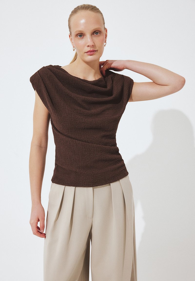 Naine, kes kannab varrukateta tekstureeritud tumepruuni pusa ja beeže volditud laiade säärega pükse, seisab valge ühevärvilise tausta ees.