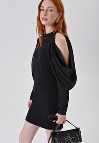 Schwarzes Kleid mit gepufften Ärmeln und ausgeschnittenem Schulterdesign. Glatte Textur, Oberschenkel-lange Länge. Schwarze Handtasche mit silbernen Akzenten.