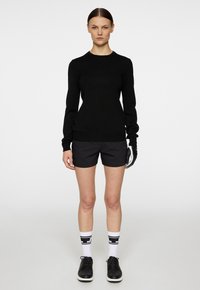 Svart långärmad tröja, grå shorts, vita strumpor med svarta ränder och svarta skor. Modell står på en enfärgad bakgrund, händerna vid sidorna.