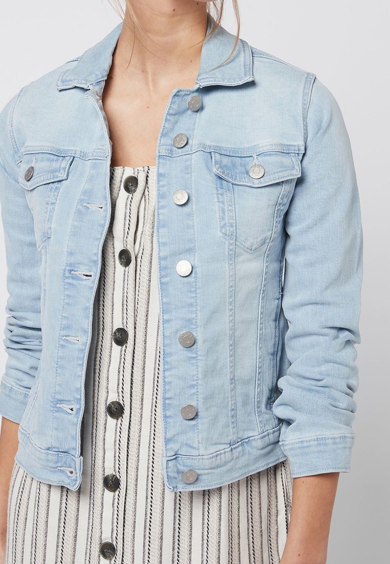 Mujer con una chaqueta de mezclilla azul claro sobre un vestido blanco con rayas verticales negras y grandes botones en la parte delantera.