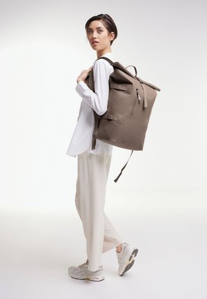 Sac à dos gris en tissu texturé, avec des bretelles ajustables, un design à rabat enroulé et une poche zippée à l'avant. Le modèle porte une chemise blanche.