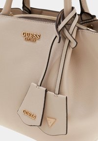 Lichtbeige leren handtas met een textuurafwerking, gouden "GUESS" logo, twee kleine labels en zwarte accenten langs de randen en handvatten.
