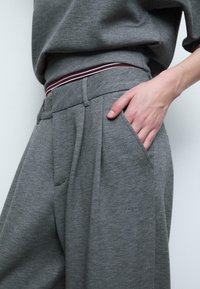 Szare spodnie dresowe z teksturowanej tkaniny, z przodu plisowane, z bocznymi kieszeniami oraz kontrastowym bordowym i białym paskiem w talii.