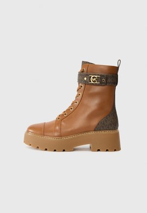 Botte de combat en cuir marron avec une semelle épaisse beige, lacets sur le devant, détails en tissu à motifs, et une sangle décorative à boucle dorée autour de la cheville.