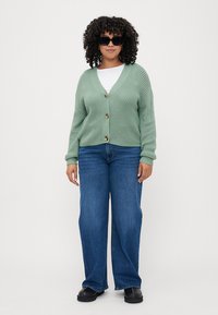 Button-up cardigan in light green knit fabric, featuring long sleeves, a V-neck, and large buttons, paired with wide-leg blue jeans.
