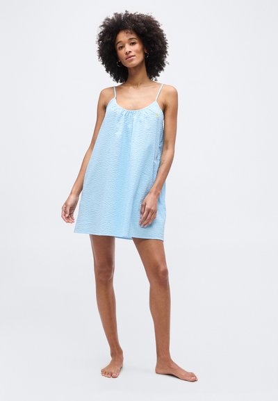 Γυναίκα με σγουρά μαλλιά που φοράει ανοιχτό μπλε, αμάνικο φόρεμα με λεπτές τιράντες, όρθια ξυπόλητη πάνω σε απλό φόντο.