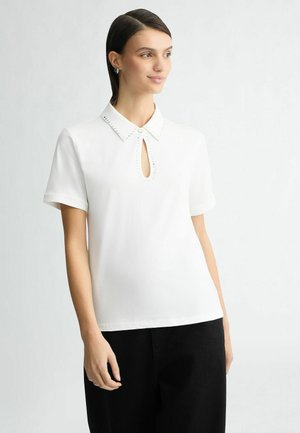 Blusa blanca de manga corta con cuello, abertura en forma de ojo de cerradura y adornos perlados a lo largo del cuello. Textura de tela suave.