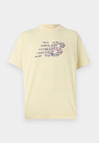 Jasnożółta koszulka z krótkim rękawem z logo New Balance składającym się z małych różowych, niebieskich i białych kształtów geometrycznych na klatce piersiowej.