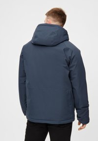 En person står med ryggen til kameraet iført en marineblå hættejakke og sorte bukser mod en simpel hvid baggrund.