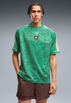 Jeune homme avec des cheveux tressés portant un t-shirt vert à motifs Puma et un short marron, debout devant un fond gris uni.