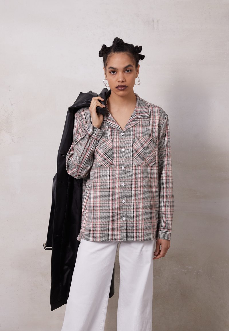 Camisa xadrez cinza e rosa com dois bolsos frontais, fecho com botão e mangas longas, combinada com calças largas brancas e um casaco preto.